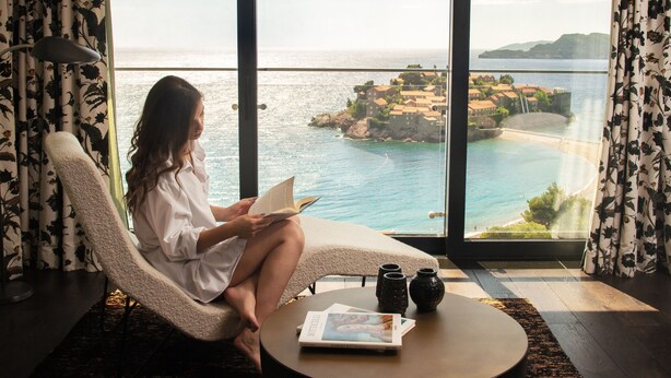 A room with a large window overlooking a coastal view, a comfortable chair, a small round table with a book and a vase, and a decorative rug. - Villa Geba Boutique Hotel (Sveti Stefan)