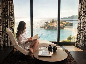 A room with a large window overlooking a coastal view, a comfortable chair, a small round table with a book and a vase, and a decorative rug.
