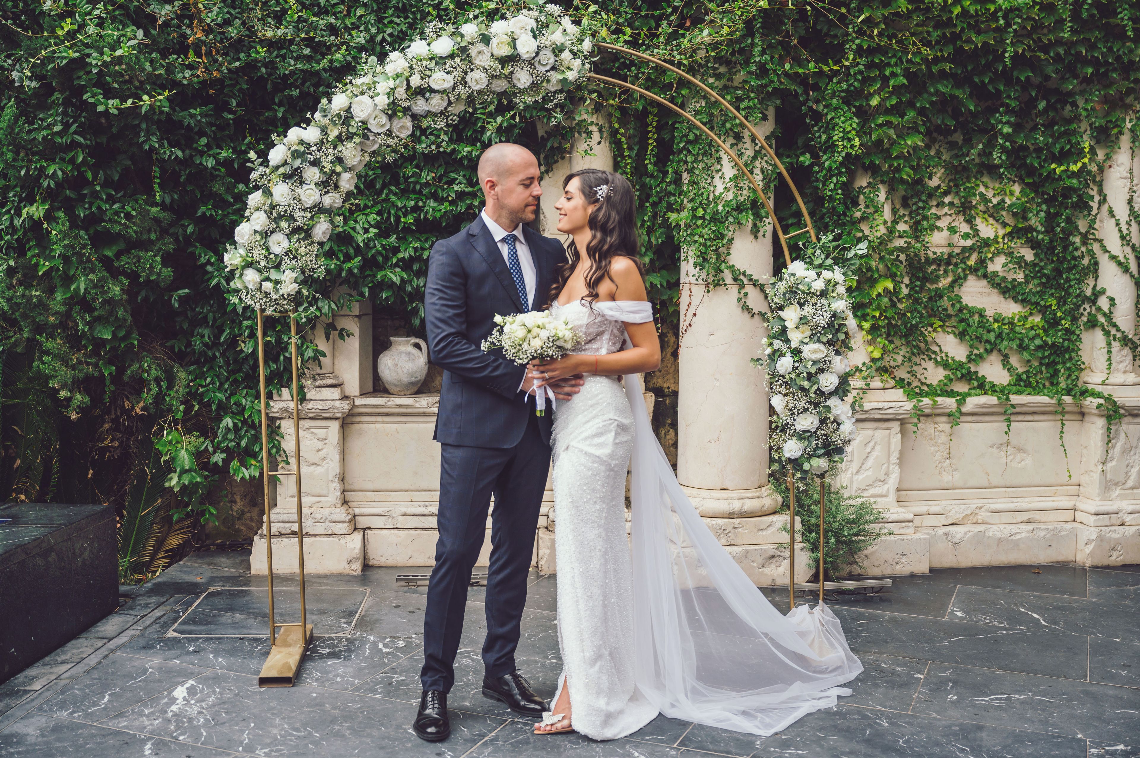 A couple standing under a floral arch with a stone wall and greenery in the background.