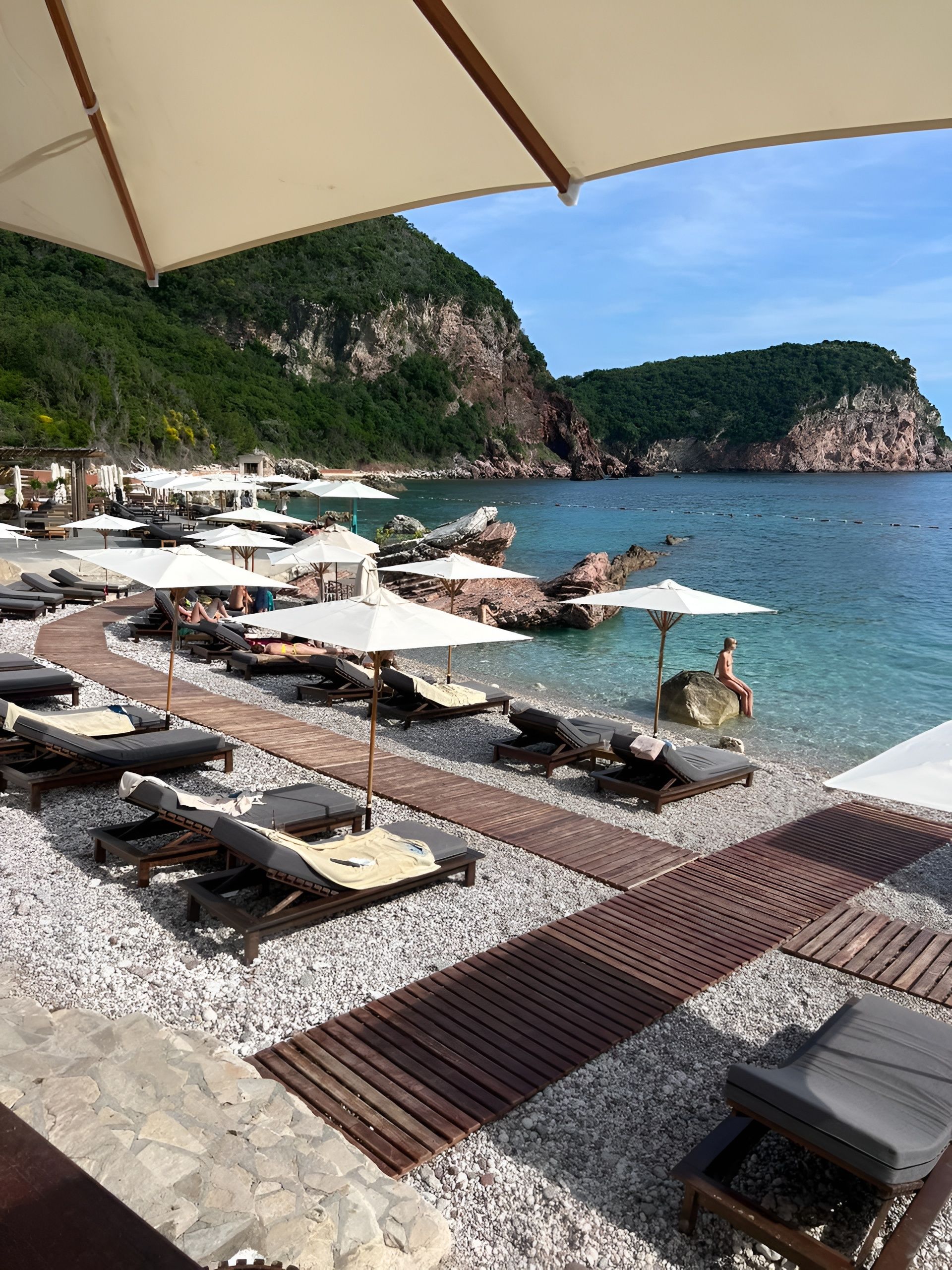 A beachside lounge area with sun loungers and umbrellas, a wooden deck, and a view of the sea and cliffs.