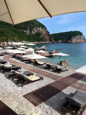A beachside lounge area with sun loungers and umbrellas, a wooden deck, and a view of the sea and cliffs. - Villa Geba Boutique Hotel (Sveti Stefan)