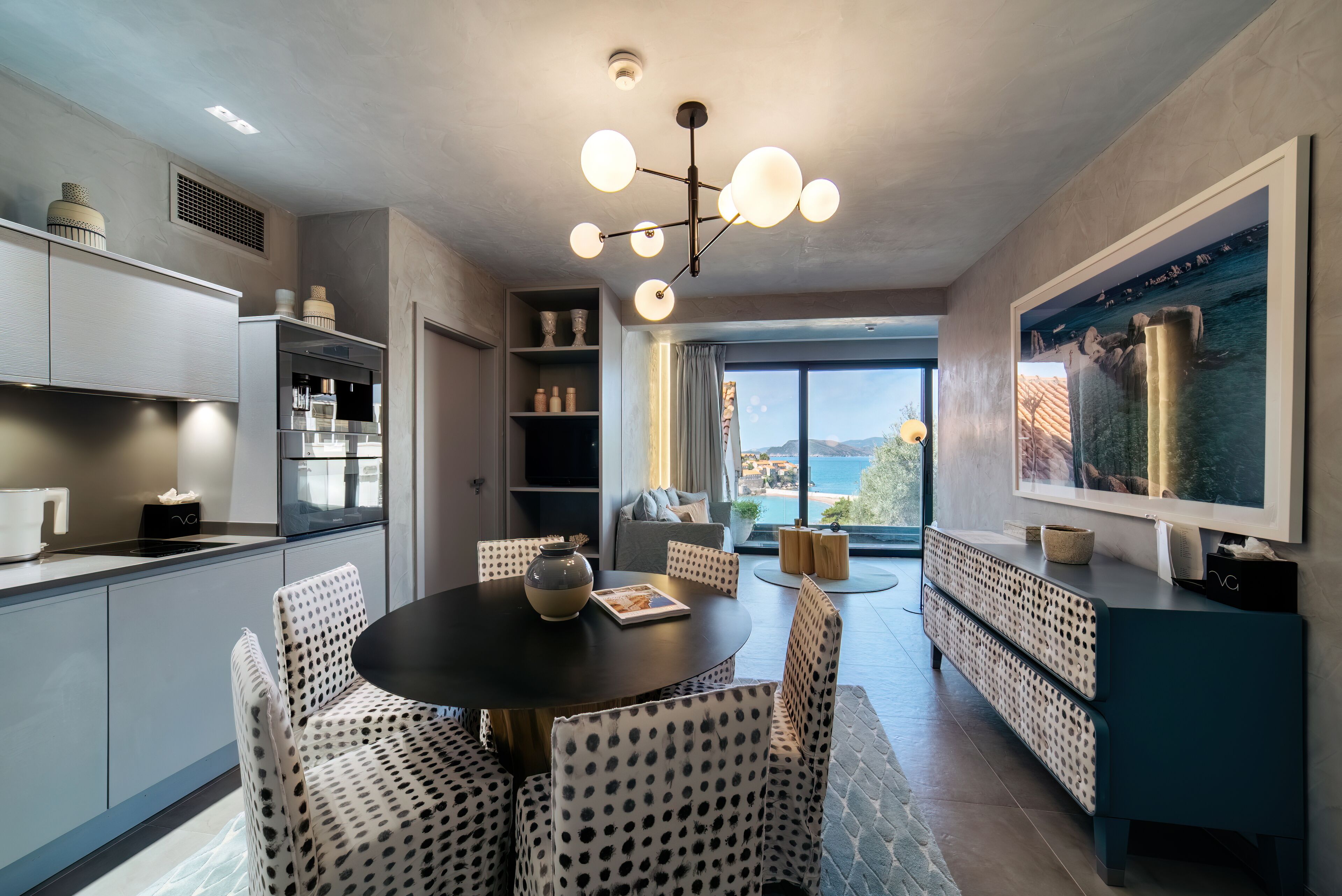 A modern kitchen with a dining area, featuring a round table with chairs, a pendant light, and a view of the outdoors.