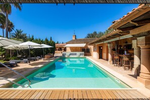 Une piscine avec des chaises longues et des parasols, un espace bar avec des chaises, et un bâtiment doté d’un toit en tuiles.