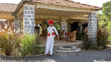 A stone building with a tiled roof, a covered porch with chairs, and a person standing with a walking stick.
