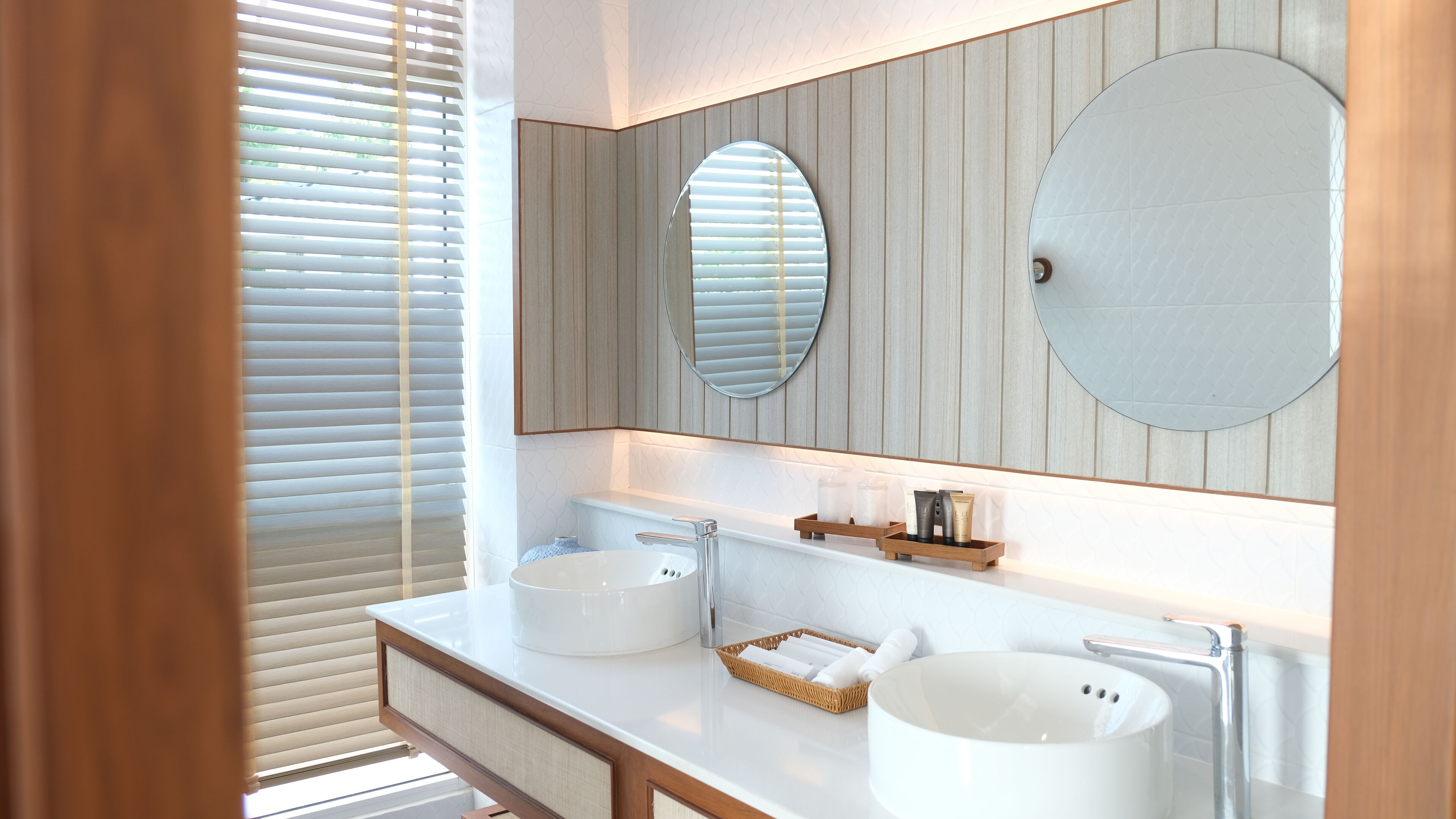 A modern bathroom with a white vanity, two sinks, a large mirror, and a wooden shelf with toiletries.