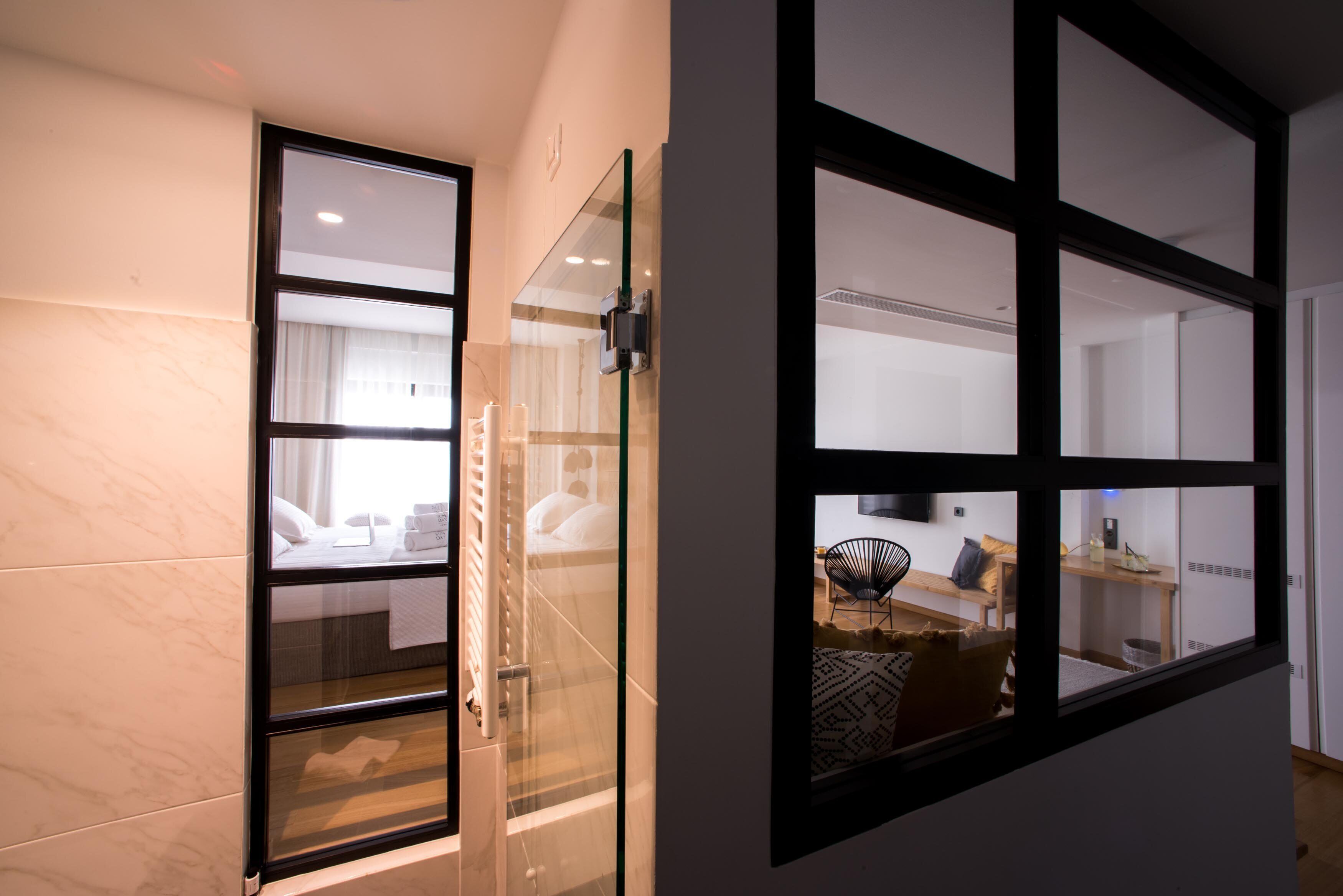 Quarto de hotel moderno com divisória de vidro, cama com lençóis brancos e vista para a sala de estar através de uma grande janela.