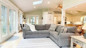 A spacious living room with a large gray sectional sofa, a wooden coffee table, and a view of the kitchen through an open door. - The Pet-Friendly Surf Shack Hideaway on the Cove (Eastham)