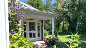 A house with a white porch, a door, and a garden with flowers and greenery. - The Pet-Friendly Surf Shack Hideaway on the Cove (Eastham)