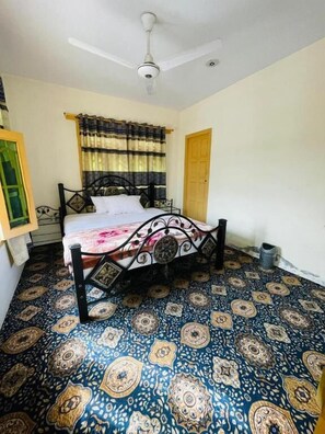 A bedroom with a bed, a ceiling fan, patterned floor tiles, a window with curtains, and a door. - Laal Haveli (Skardu)
