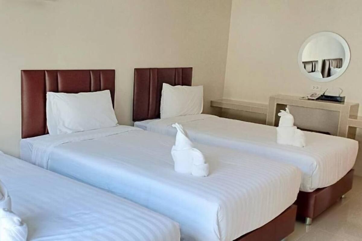 A hotel room with three beds, each with a brown headboard and white bedding. There is a round mirror on the wall and a small table with a phone and a bottle.