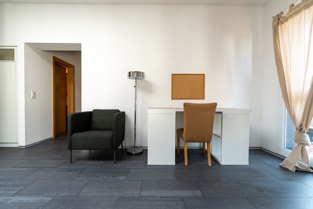 Una habitación moderna con escritorio, dos sillas, una lámpara de pie y un tablón de corcho en la pared.