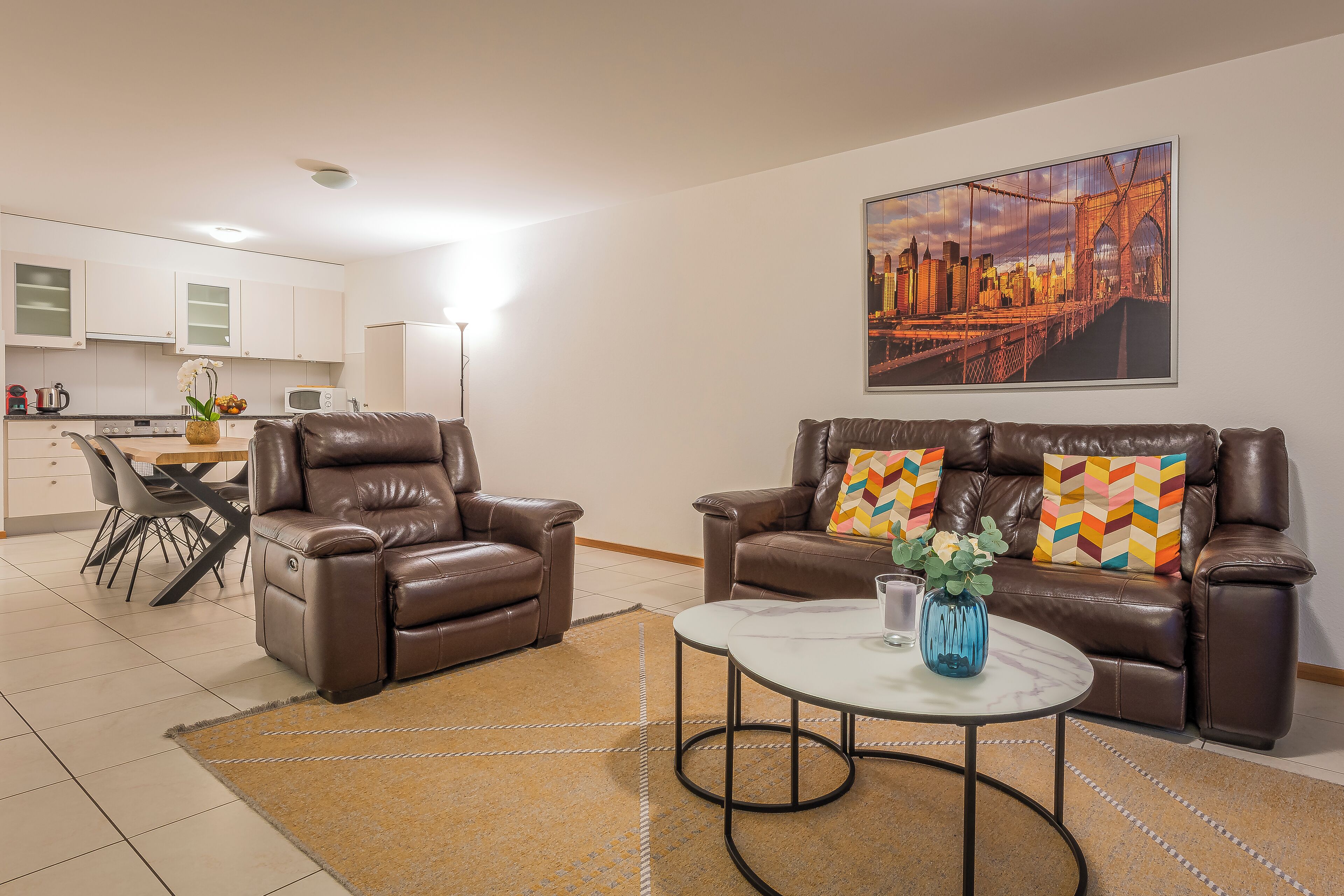 A living room with a brown leather sofa, a white coffee table, and a cityscape painting on the wall.