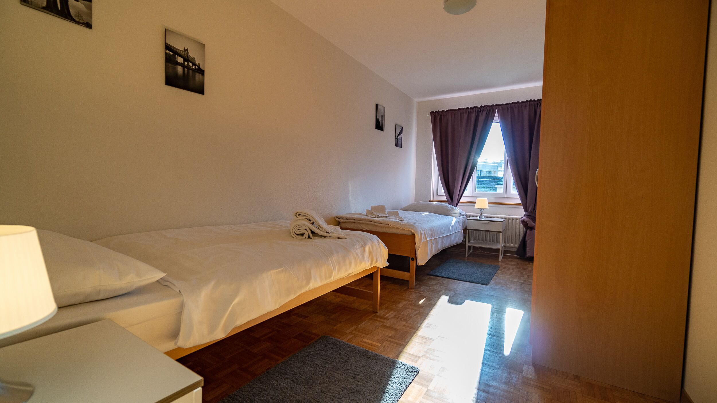 Habitación de hotel con dos camas individuales, un escritorio pequeño con lámpara, una ventana con cortinas y un radiador.