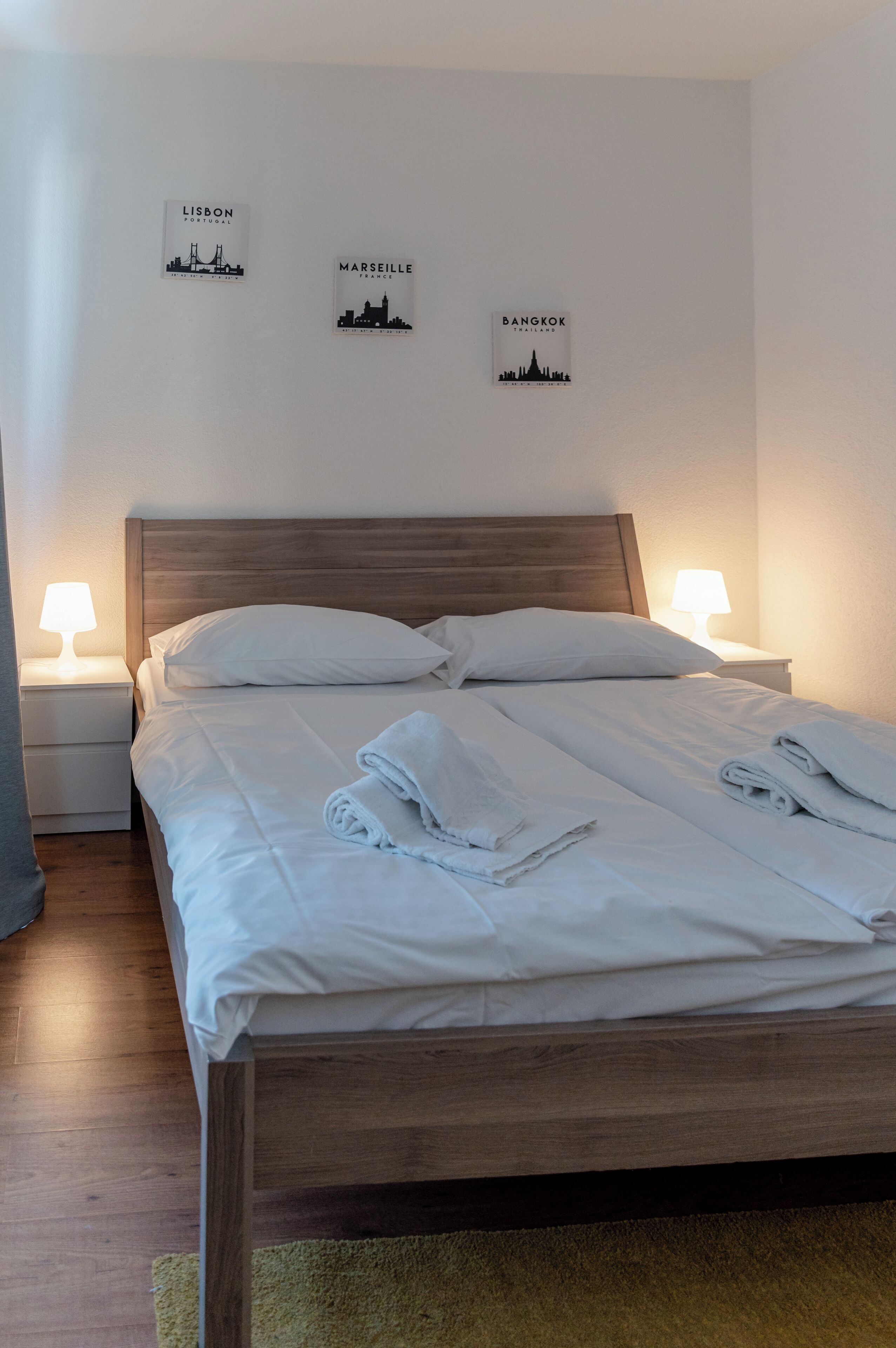 Una cama bien hecha con sábanas blancas y toallas enrolladas, dos lámparas de noche y una obra de arte con vistas de la ciudad.