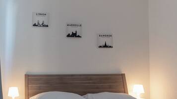 Una cama bien hecha con sábanas blancas y toallas enrolladas, dos lámparas de noche y una obra de arte con vistas de la ciudad.