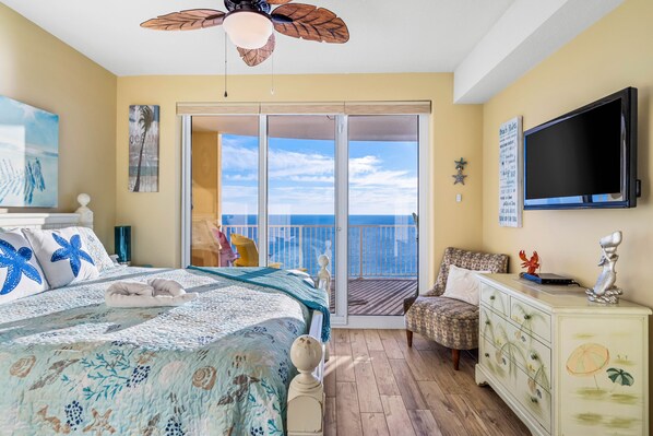 A bedroom with a bed, a dresser, a television, and a balcony with a view of the ocean.