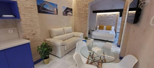 Cozy living space with a sofa, coffee table, and potted plant, featuring a comfortable bed in the background.