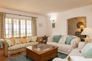 A living room with a wicker sofa, a glass coffee table, and a white sofa. There are curtains on the windows, a lamp, and a small table with a globe.