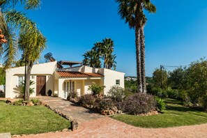 A single-story house with a tiled roof, a paved driveway, and a well-maintained garden with palm trees and shrubs. - Villa Bandicoot, Lagoa de Momprolé, Algarve (Loulé)