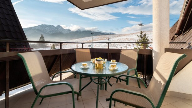 A balcony with a round table set for tea, surrounded by green chairs, overlooking a snowy mountain landscape.