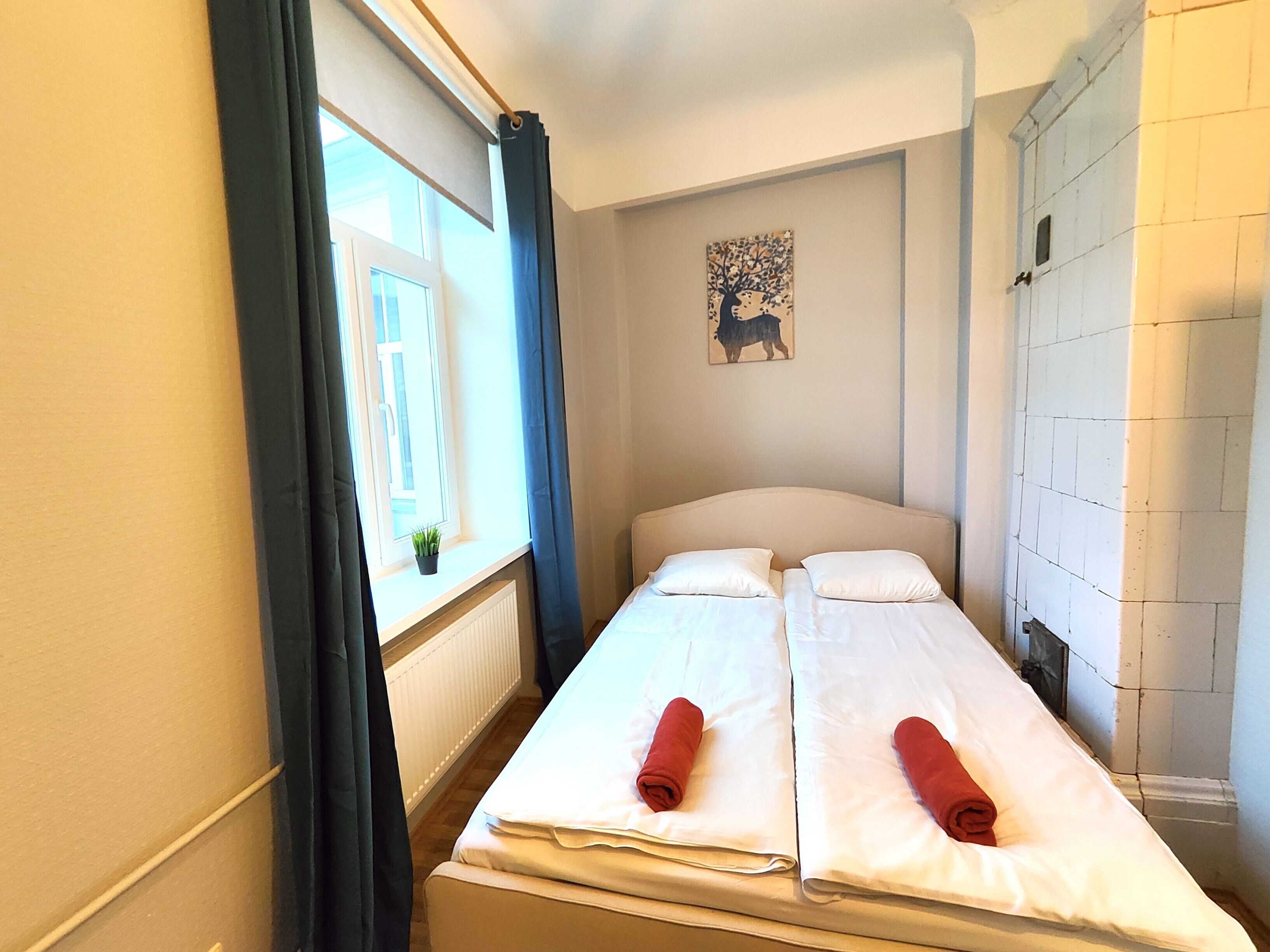 Ein kleines Zimmer mit einem Einzelbett, einem Fenster, einem Heizkörper, einem Wandbild und einem weißen Kleiderschrank.