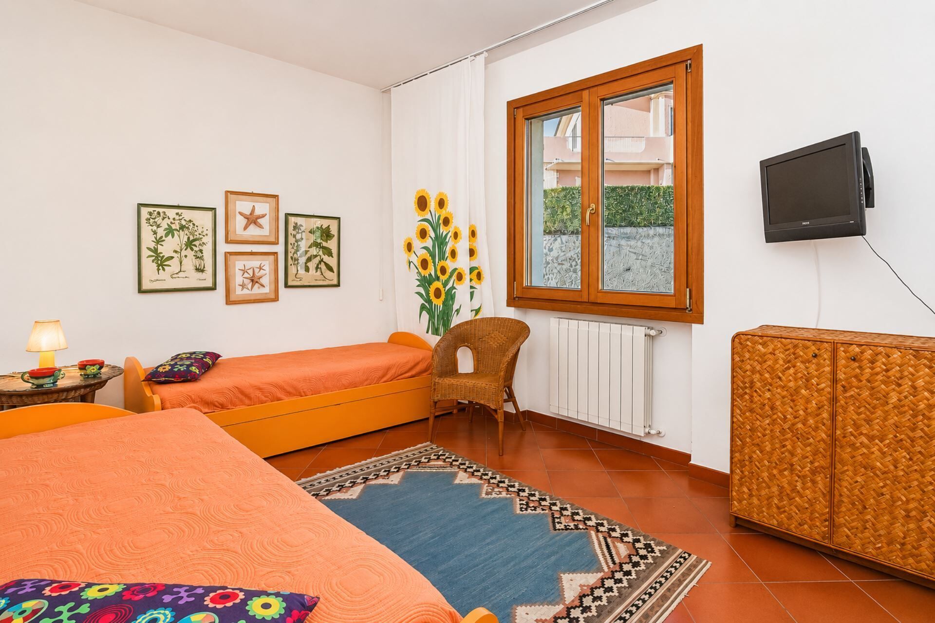 A bedroom with a bed, a chair, a small table with a lamp, a TV, and framed botanical prints on the wall.