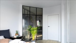 Un dormitorio moderno con un gran ventanal corredera, una cama con ropa de cama blanca, una mesita de noche con una planta y una puerta blanca.