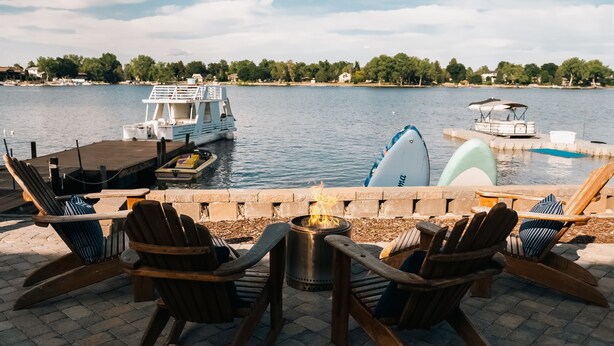Eine Seeterrasse mit Adirondack-Stühlen, einem Feuerkorb und Booten im Hintergrund.