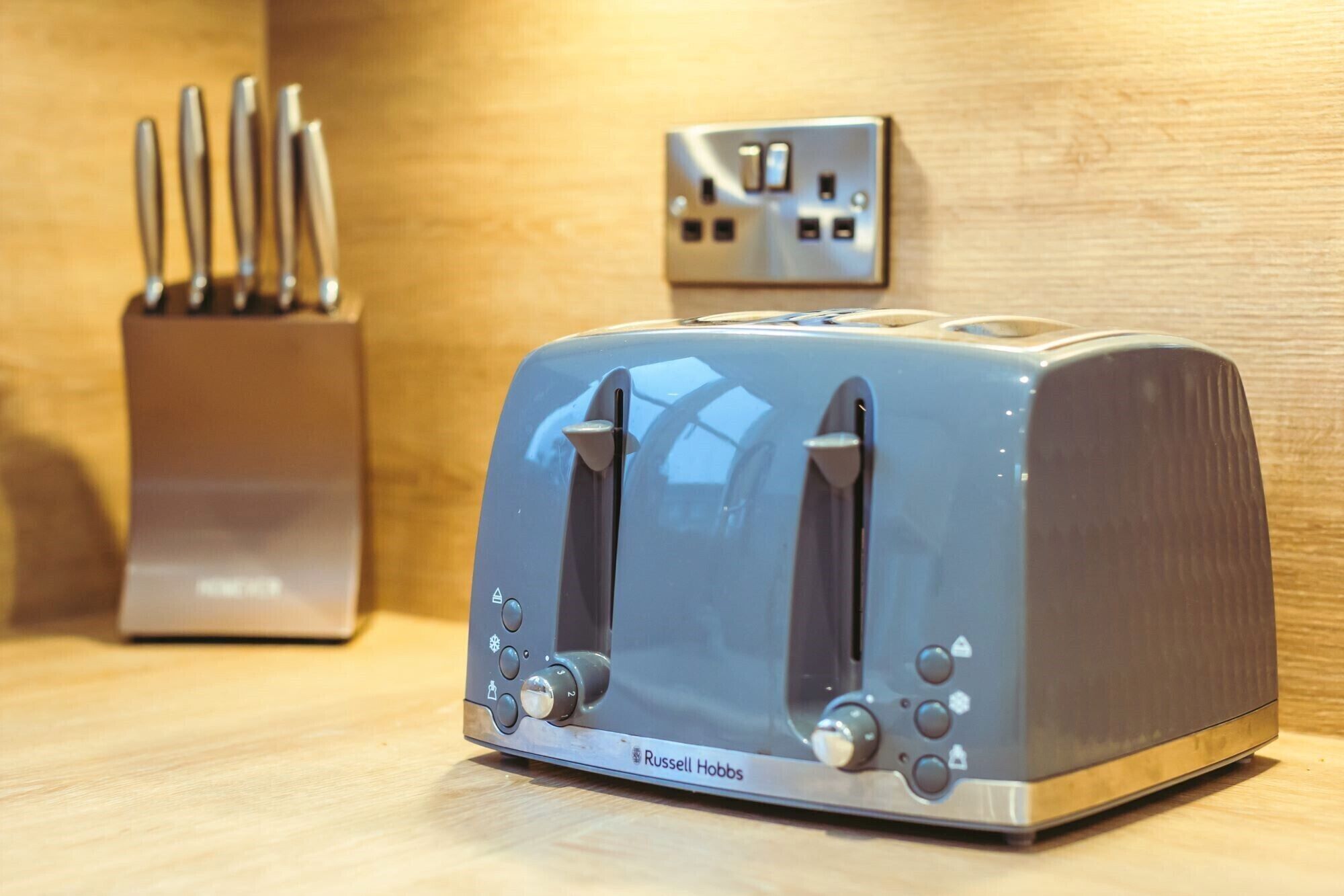 Un tostador Russell Hobbs sobre una encimera de madera, con un bloque para cuchillos y un enchufe de corriente al fondo.