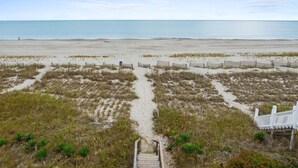Vista sulla spiaggia con una scala in legno che scende sulla sabbia, una recinzione bianca e un cielo sereno.
