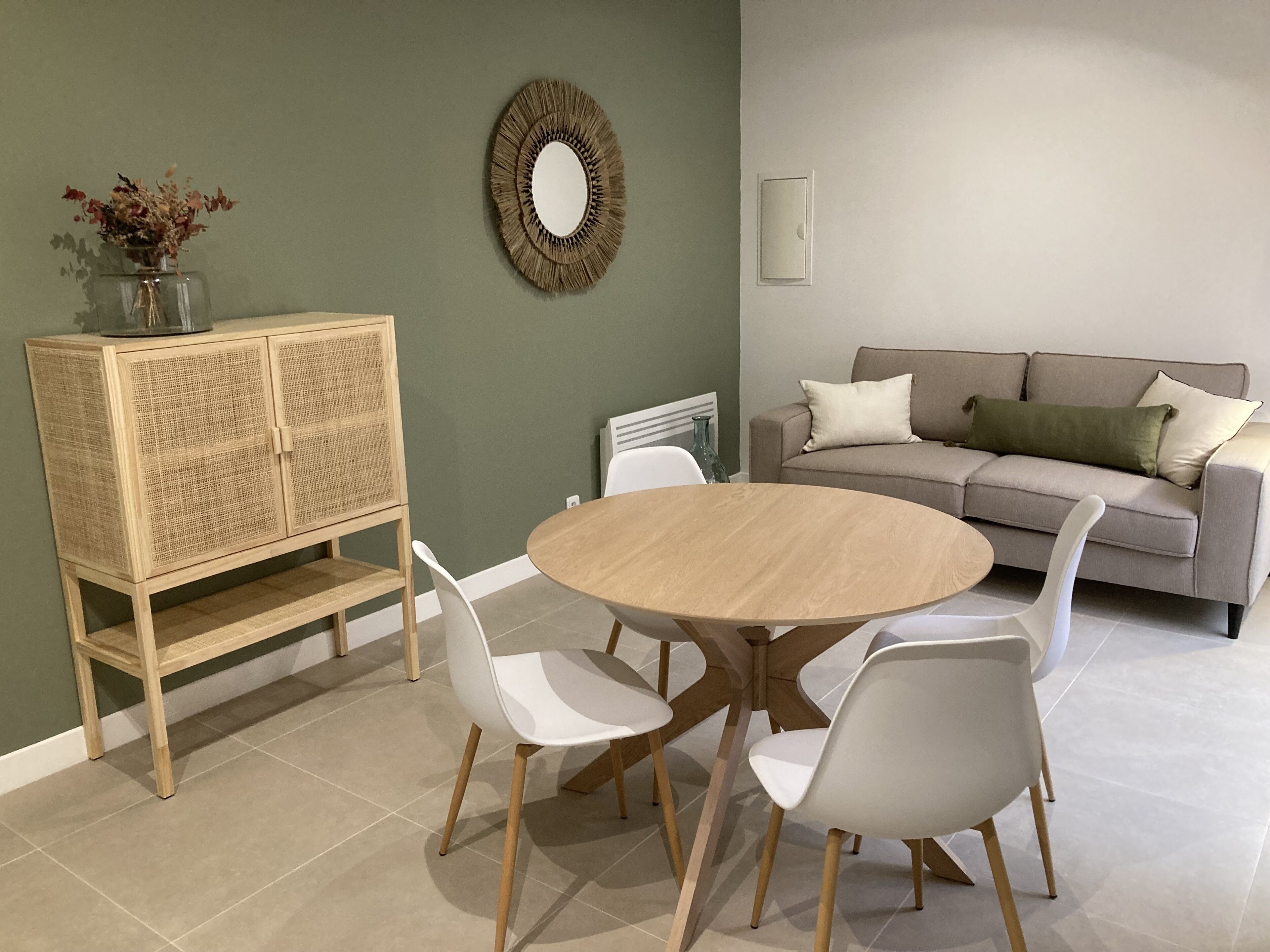 Un coin repas avec une table ronde en bois entourée de chaises blanches, un meuble en osier, un canapé gris avec des coussins et un miroir fixé au mur.