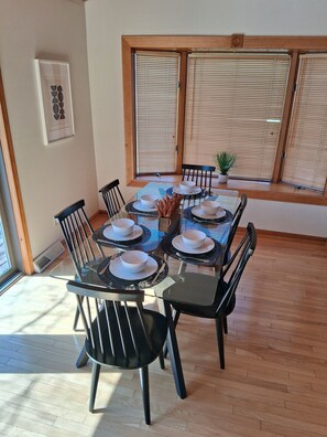 Un coin repas avec une table dressée pour quatre personnes, entourée de chaises, avec une fenêtre et des stores en arrière-plan.