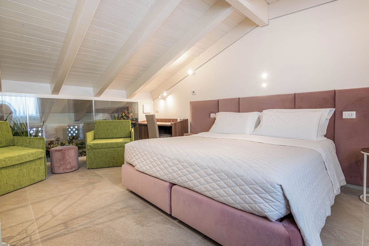 Quarto moderno com uma cama grande, poltronas verdes e um banquinho rosa.