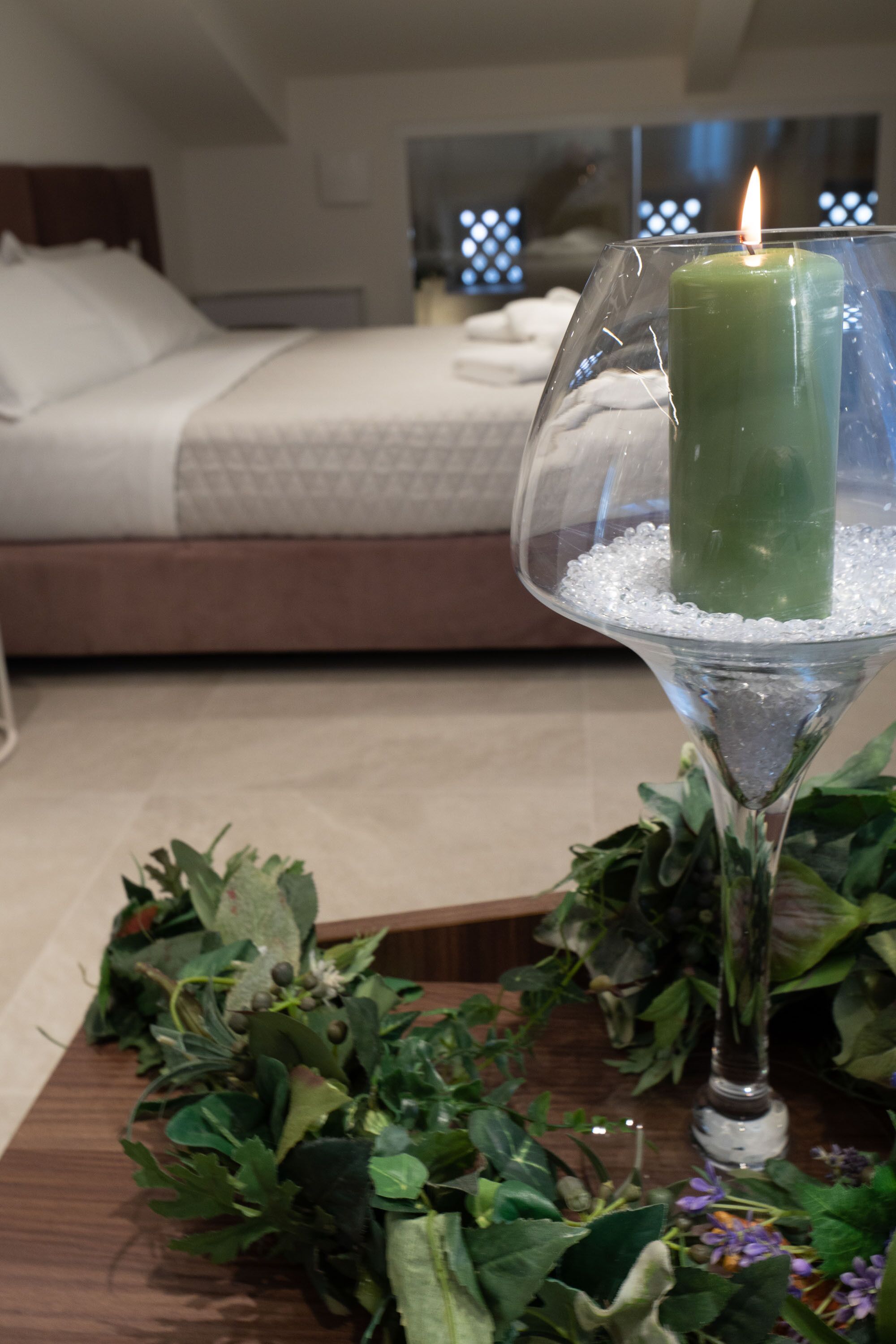 A room with a bed, a green candle in a glass holder, and a table with greenery.