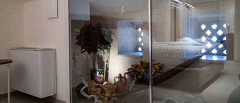 Una habitación moderna con una puerta de vidrio corredera que da a un dormitorio. Hay una mesita con una silla, una planta en maceta y un adorno en el suelo.
