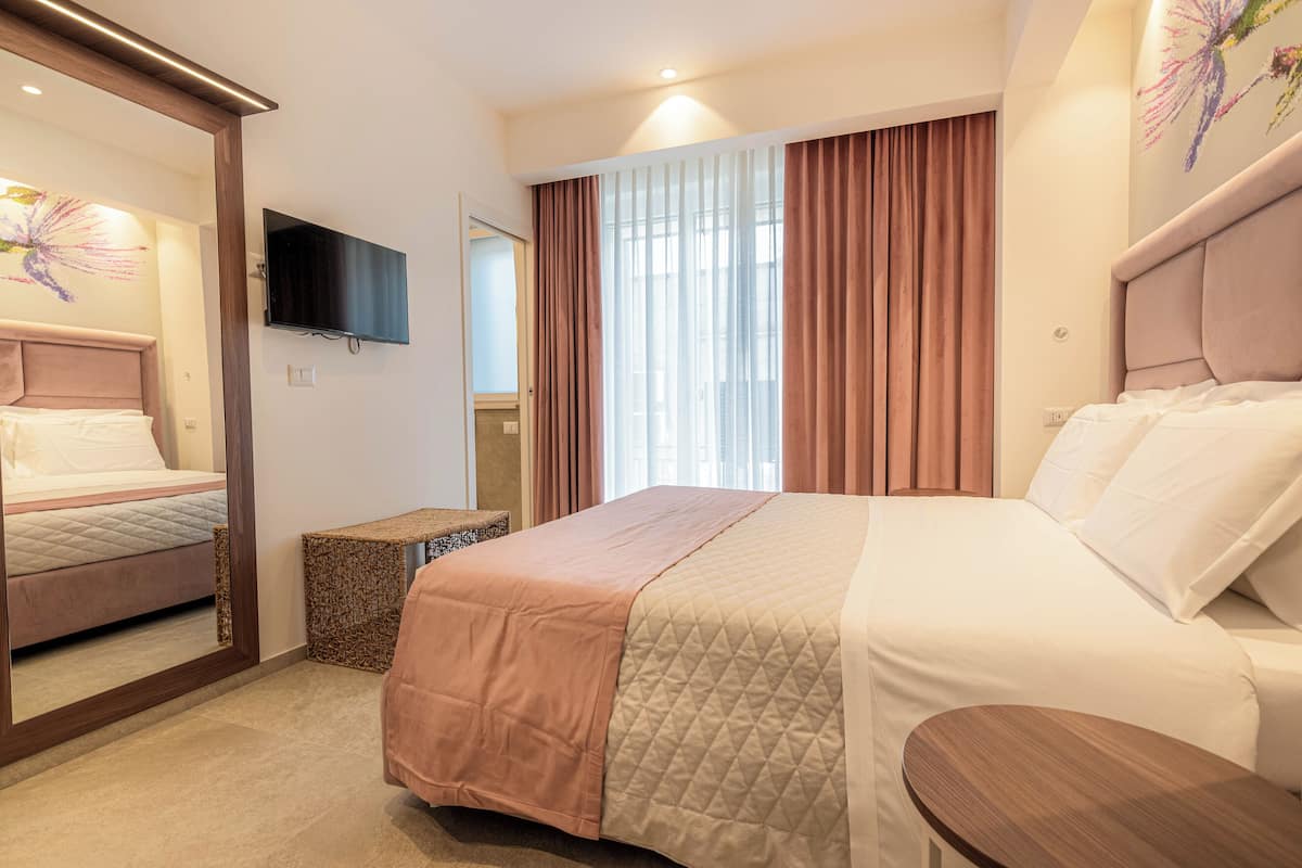 Quarto de hotel com uma cama grande, televisão na parede, espelho e janela com cortinas.