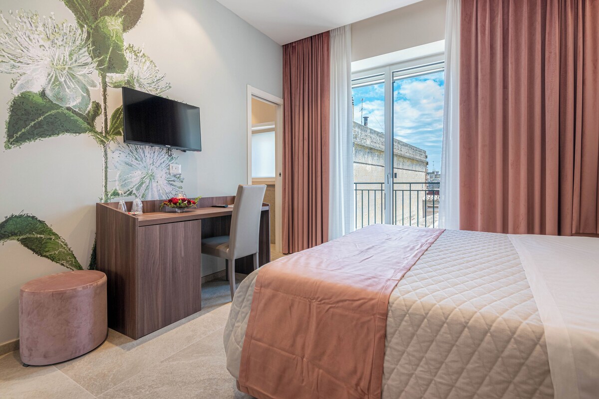 Hotellihuone, jossa on sänky, työpöytä, tuoli, televisio ja suuri ikkuna, jossa on verhot.