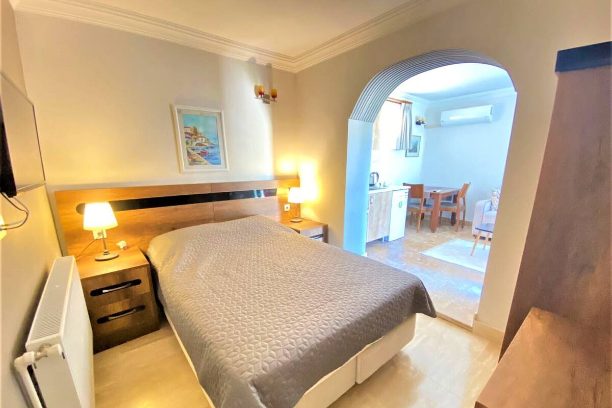 Una camera d'albergo con un letto, abat-jour, un termosifone, un quadro appeso al muro e un'apertura che dà su un'area ristoro.