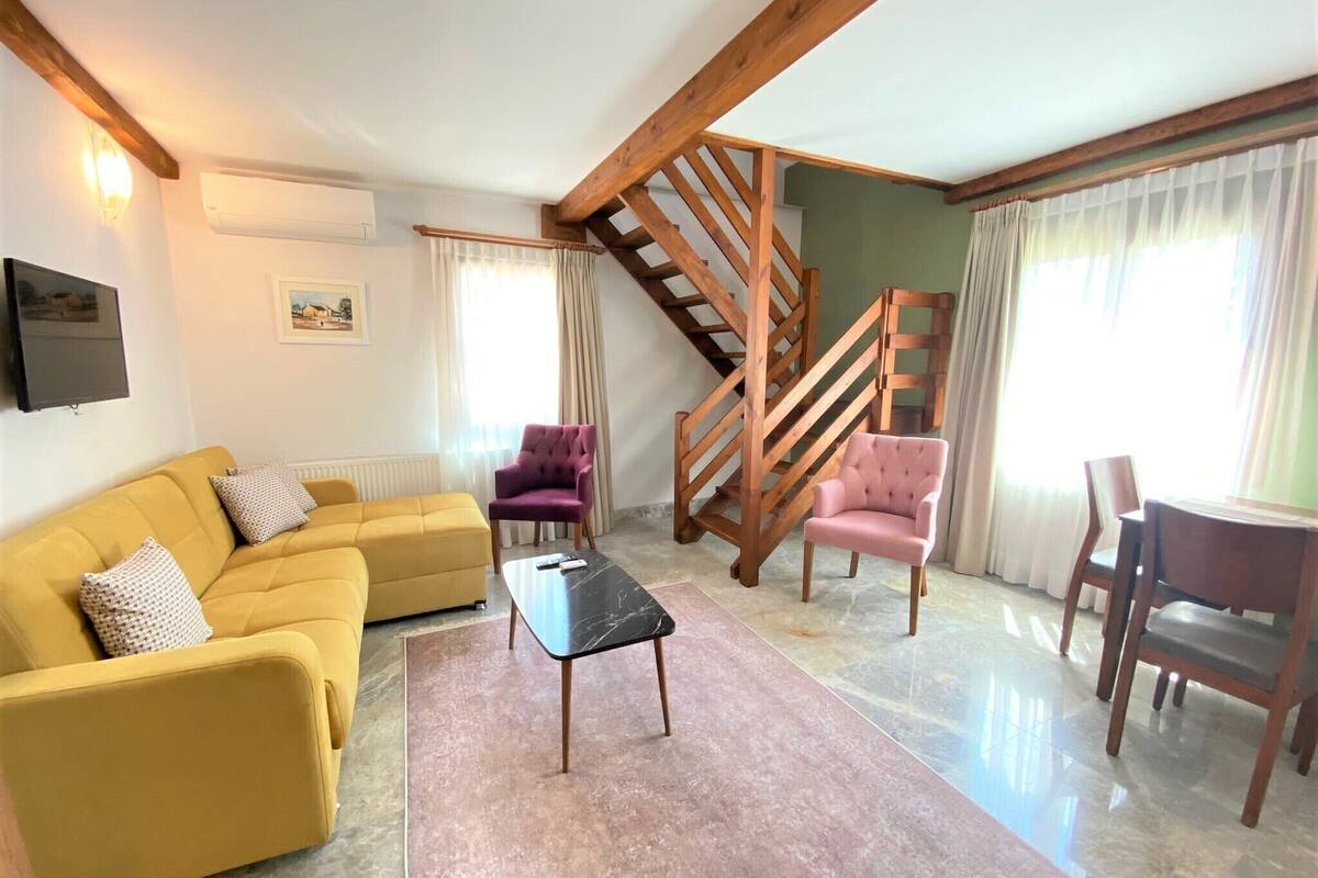 Un soggiorno con un divano giallo, una poltrona rosa, una scala in legno e un'area pranzo con tavolo e sedie.