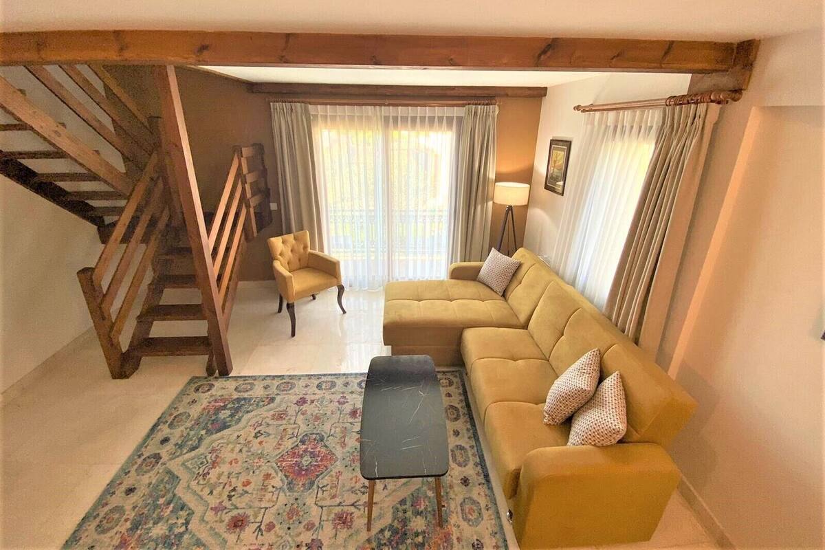 Un soggiorno con divano, poltrona e un piccolo tavolino. Si vede una scala con ringhiere in legno che conduce a un piano superiore.