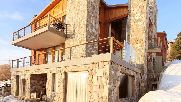 A modern two-story house with stone walls, a balcony, and a hot tub in the foreground.