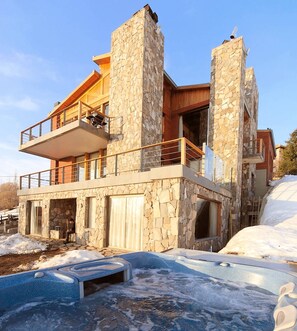 Una casa moderna de dos pisos con paredes de piedra, un balcón y un jacuzzi en primer plano.