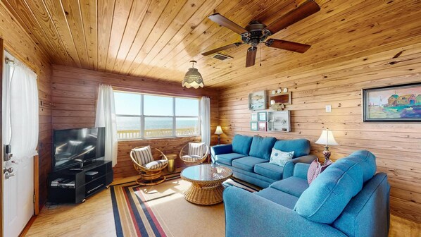 Un interior de cabaña acogedor con un sofá azul, sillones de mimbre, un televisor y un ventilador de techo.