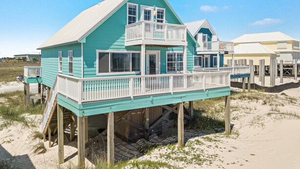Ein türkisfarbenes Strandhaus mit Balkon, Holzterrasse und Sandstrand.