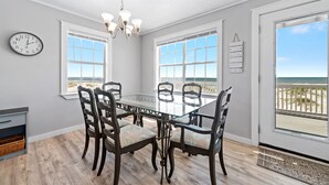 Ein Esszimmer mit Glastisch und Stühlen, großen Fenstern mit Blick auf den Strand, einer Kristallleuchter und einer Wanduhr.