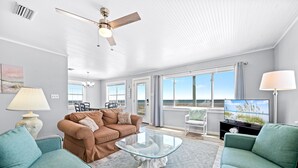 Ein modernes Wohnzimmer mit einem Glastisch, zwei Sofas, einem Deckenventilator und großen Fenstern mit Blick ins Freie.