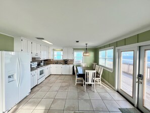 A kitchen with white cabinets, a dining table with chairs, a refrigerator, and a view of the ocean through large windows.
