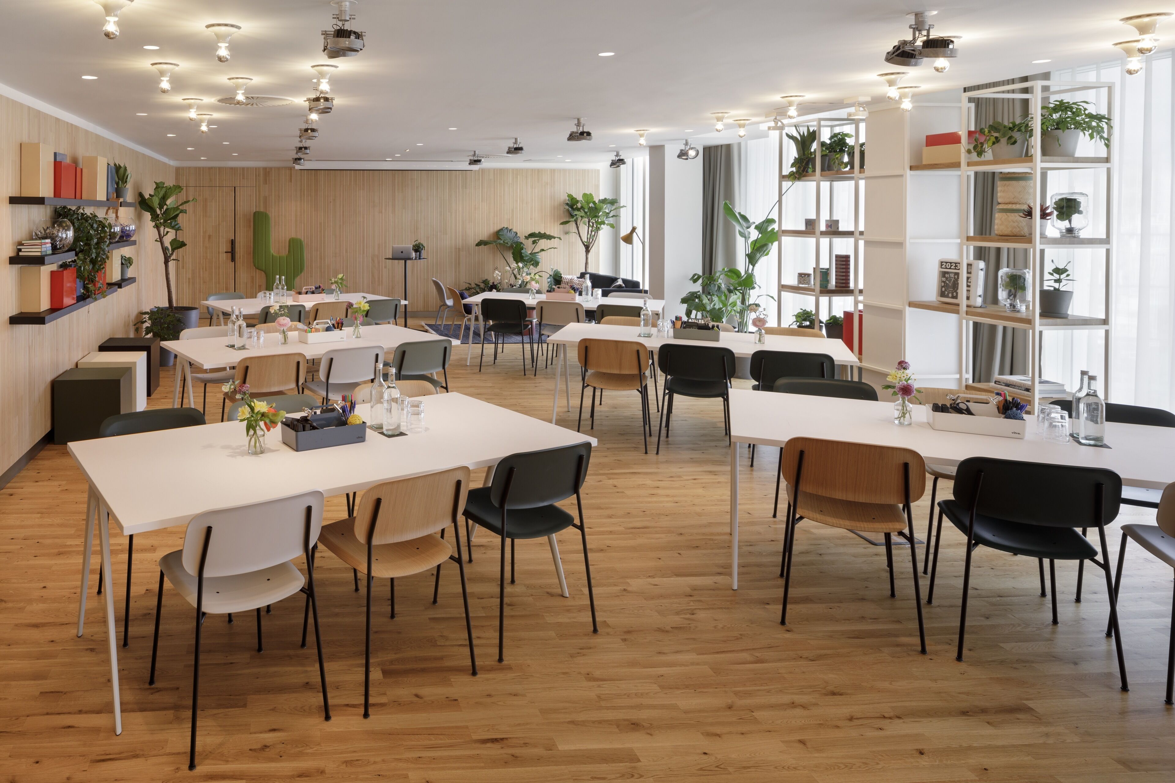 Un espace repas moderne doté de plusieurs tables et chaises, d’un plancher en bois et d’étagères ornées de plantes et de livres.