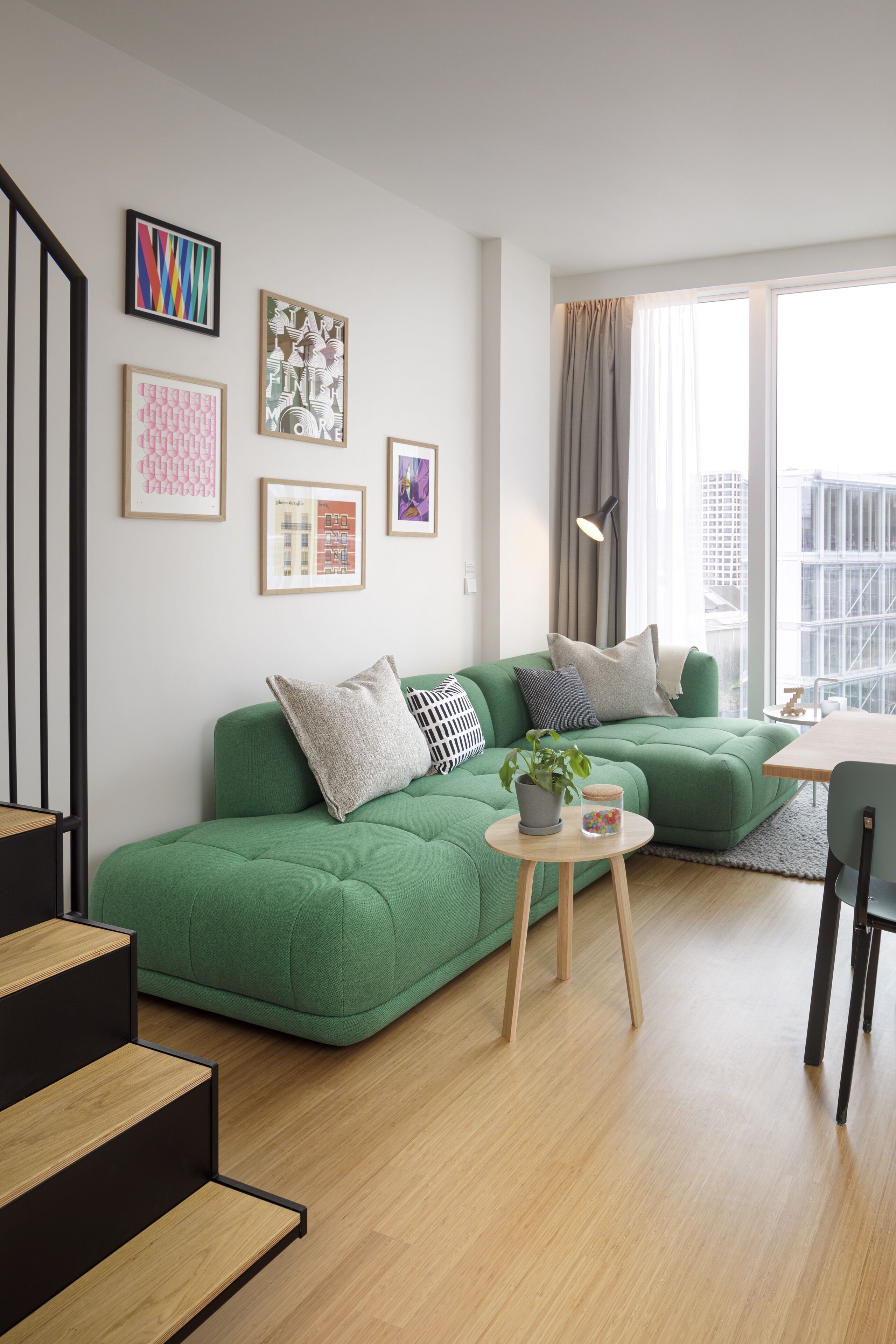 A modern living room with a green sofa, a small round table with a plant and a jar, and a staircase with black railings.