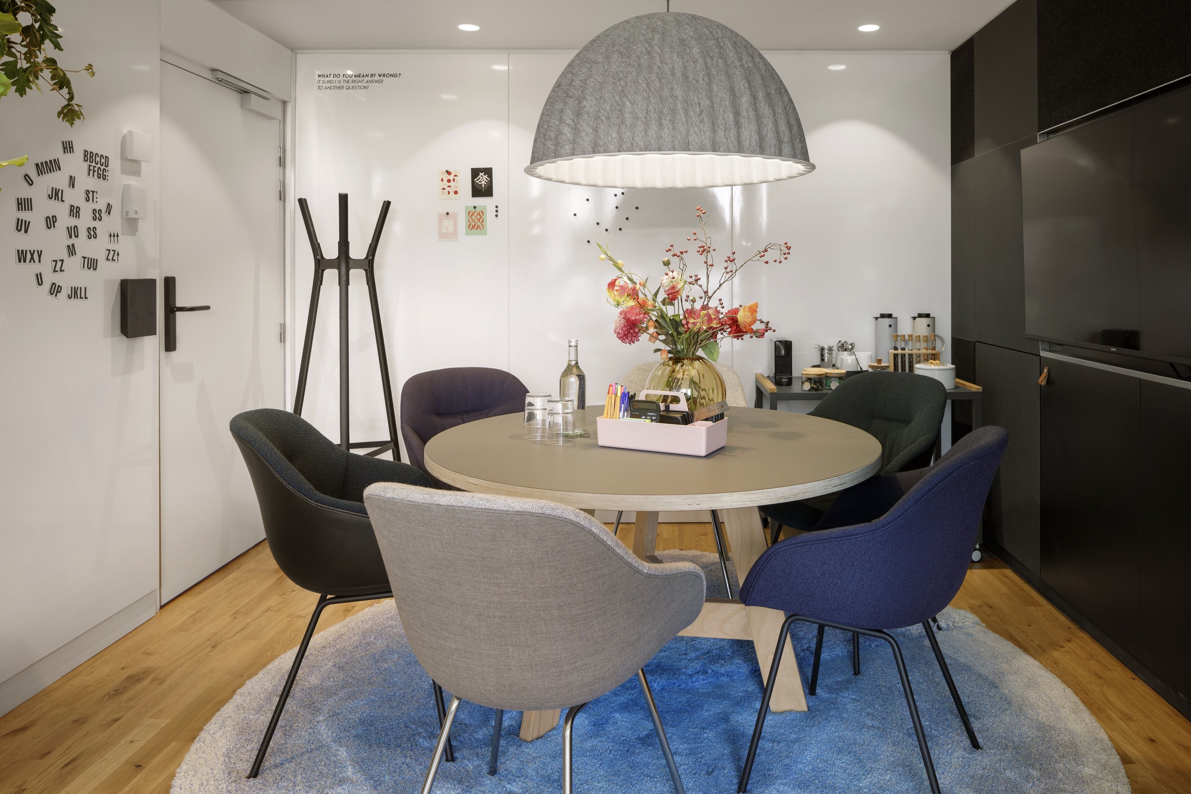 A modern dining area with a round table, four chairs, a central vase with flowers, a large pendant light, and a wall-mounted television.